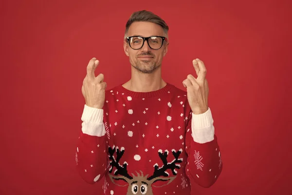 making a wish crossing fingers. happy man in winter sweater and glasses. xmas guy on red background. happy new year. merry christmas.