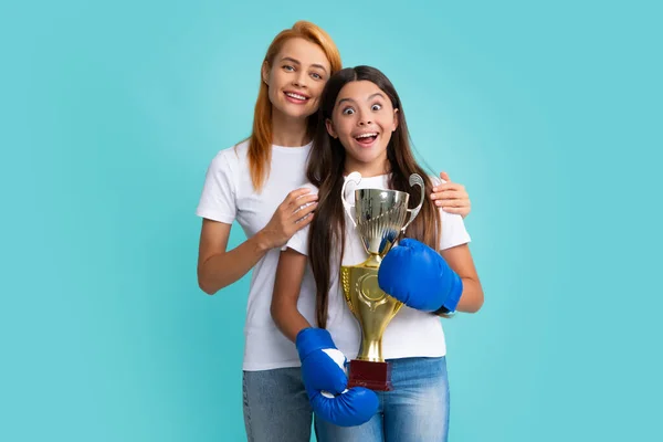 Strong family, girl power. Sportive young woman and her daughter wearing boxing gloves, holding winning prize, showing trophy, emotional excited and happy
