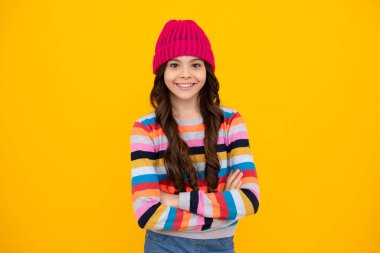 Beautiful teen girl in a winter hat and a warm sweater. A child on a yellow isolated background. Happy teenager, positive and smiling emotions of teen girl