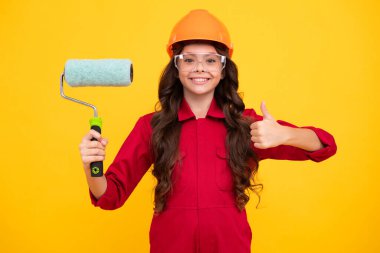Kids renovation and building concept. Little girl in protective helmet and paint brush for painting walls isolated on yellow background. Happy face, positive and smiling emotions of teenager girl