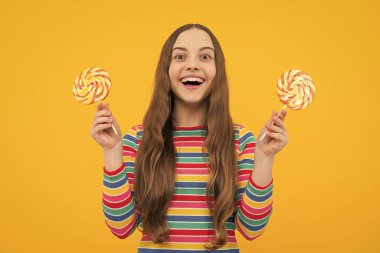 Şekerli lolipop yiyen genç bir kız. Çocuklar için şeker ve şeker. Çocuk, sarı izole arka planda lolipop dondurması yiyor. Lezzetli karamel, şekerci