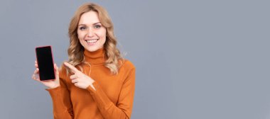 Kadın portresi, kopyalanmış başlık pankartı. Kadın boş cep telefonu ekranını işaret ediyor. Gri arka plan fotokopi alanı, akıllı telefon reklamı.