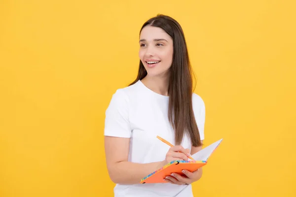 Smiling young woman college student holding book on isolated yellow background. Model emotionally showing facial expressions. Portrait of a pensive young girl making note