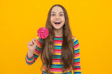 Funny child with lollipop over yellow isolated background. Sweet childhood life. Teen girl with yummy caramel lollipop, candy shop. Teenager with sweet sucker