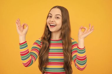 Küçük kız sarı arka planda izole edilmiş işaret dilini gösteriyor. Komik ergen yüzü.