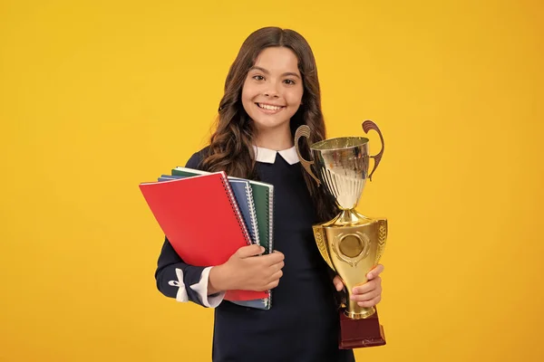 Schoolgirl in school uniform celebrating victory with trophy. Teen holding winning award prize on yellow background. Child hold books with gold trophy or winning cup. Education graduation, victory