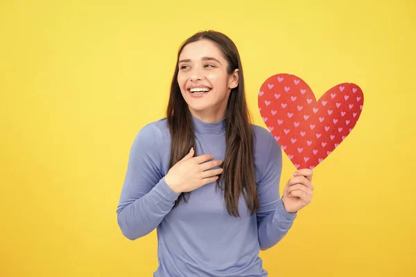 Girl feeling love. Beautiful girl holding valentines gift on yellow background. Portrait of young woman holding red paper heart