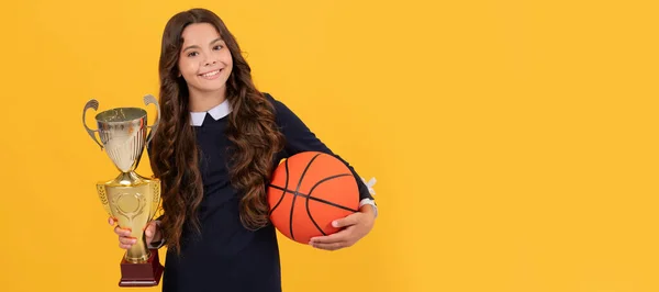 幸せな10代の女の子は黄色の背景にバスケットボールボールとチャンピオンカップを保持します チャンピオン 孤立した子供の顔 バナーヘッダー コピースペースの水平ポスター — ストック写真