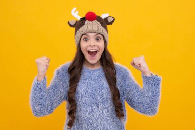 Güzel kış çocukları portresi. Kış süveteri giyen ve sarı arka planda şapka ören genç kız. Heyecanlı bir yüz, neşeli genç bir kız.