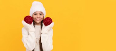 Kışlık şapkalı ve eldivenli neşeli çocuk sarı arka planda dekoratif xmas topları tutuyor, xmas. Noel çocuğu, stüdyo çocuğu kışın fotokopi çekilen portresi