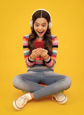 Cep telefonu tutan hayret verici ergen çocuk. Güzel bir genç kızın cep telefonu, cep telefonu uygulaması kullanarak yakın plan portresi. Parlak canlı sarı renkli arka planda izole edilmiş. Heyecanlı genç kız