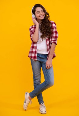 Teenager child girl in headphones listening music, wearing stylish casual outfit isolated over yellow background. Happy teenager with earphones, positive and smiling emotions of teen girl