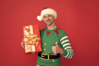 Mutlu noel hediyeleri. Başparmağını kaldır. Elf kostümü ve Noel Baba şapkası giymiş mutlu bir adam. Kırmızı arka planda hediye kutusu olan xmas adamı. Mutlu yıllar..