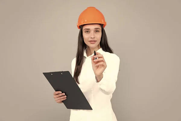 Young woman in hard hat helmet with clipboard isolated on grey background. Architect woman wear helmet and shirt. Business woman, manager, builder. Female worker in hardhat helmet pointing you