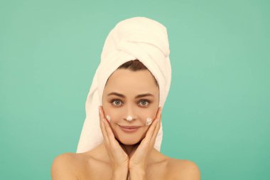 Gülümseyen genç bir kadın mavi arka plana yüz kremi sürüyor..