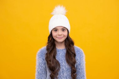 Güzel kış çocukları portresi. Kış süveteri giyen ve sarı arka planda şapka ören genç kız. Mutlu kız yüzü, pozitif ve gülümseyen duygular