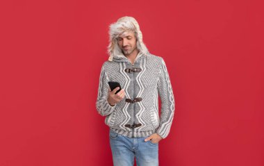 Kazaklı ve kulaklıklı mutlu adam kırmızı arka planda telefondaki e-postayı kontrol ediyor..