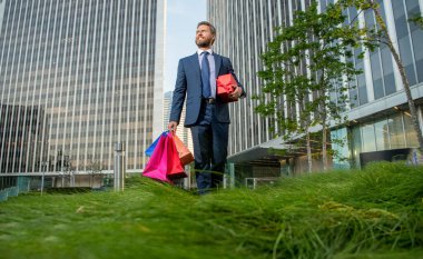 Gülümseyen, resmi giyinen, alışveriş çantalı ve hediye kutusuyla ofisin dışında yürüyen başarılı bir adam..