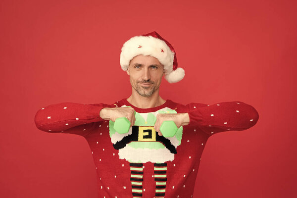 Счастливый Санта-мужчина улыбается, делая гантели тренировки на красном фоне, Рождество.
