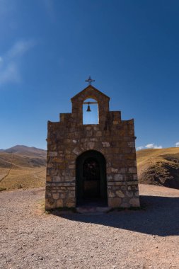 Güzel bir günde, dağlarda tek başına küçük bir şapel. Bazı yollar arkada görülebilir. Şapel farklı boyutlarda taşlarla yapılır. Üst tarafında bir çan ve bir haç var.
