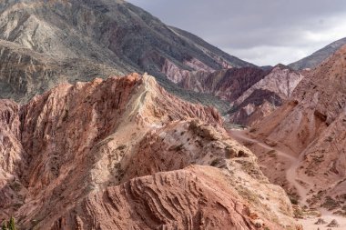 Kırmızımsı tonların arasındaki toprak yol. Bir vadi ve dağlar çeşitli renklerdedir. Bir vadi boyunca dağların manzarası.