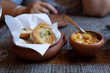 İki kil tencereli bir kadın eli masada, biri tostlu, diğeri de soğan ve fasulyeli. Masa tahtadan yapılmıştır ve kırsal bir masa örtüsü vardır..