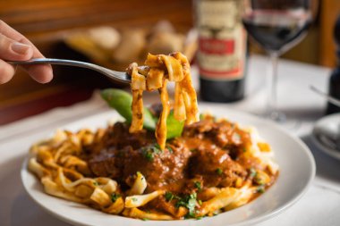 Kırmızı soslu, kurdeleli erişteli çatal tutan bir elin yakın çekimi. Bir restoranda, masada bir bardak şarap ve ekmek sepetiyle..