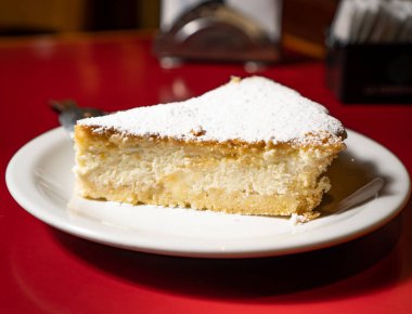 Kırmızı bir masada beyaz bir tabakta, üzerinde krema şekeri olan ricotta kekinin ayrı bir kısmı..