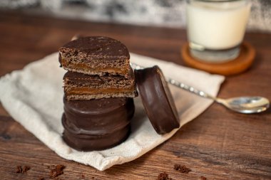a close-up of a stack of several chocolate cookies on a wooden table, with a black and white textured background, behind it is a glass cup with milk