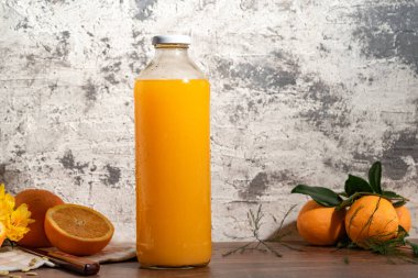 a glass bottle with orange juice, leaning on a wooden table, there are also whole oranges and another one cut in half. next to it you can see a knife and some yellow flowers.