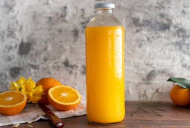 a glass bottle with orange juice, leaning on a wooden table, there are also whole oranges and another one cut in half. next to it you can see a knife and some yellow flowers.