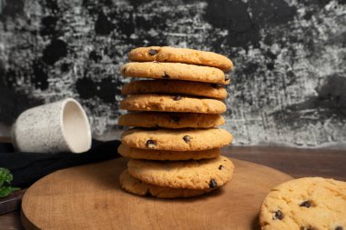 a stack of chocolate chip cookies. on a wooden board and behind you can see a cup. the background is black and white, textured.