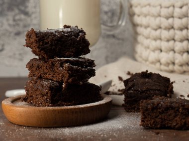 close up of a stack of brownies. with a square shape, they are on a wooden board. behind you can see a cup with milk and a container with several eggs.