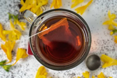 closeup and top view of a glass cup with tea. you can see the tea bag. there are yellow petals around the cup