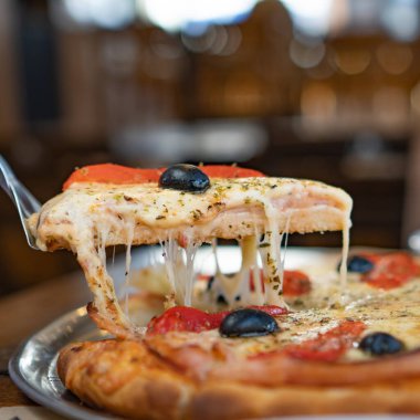 a spatula is seen being held, grasping a slice of pizza. the pizza is ham, bell pepper and cheese with black olives.