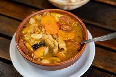 a pot with tripe, vegetables, sauce is observed. everything is served hot. There is also a few breads behind on the wooden table.