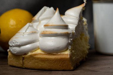 a slice of cake served on a wooden board. the cake is lemon pie, the meringue is in the form of cones on top of the delicious yellow lemon cream. there are also some lemons