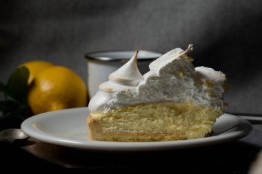 a portion of lemon pie, with lemon-flavored yellow cream and meringue cones on top. delicious cake serving on a white plate. there are lemons and a cup.