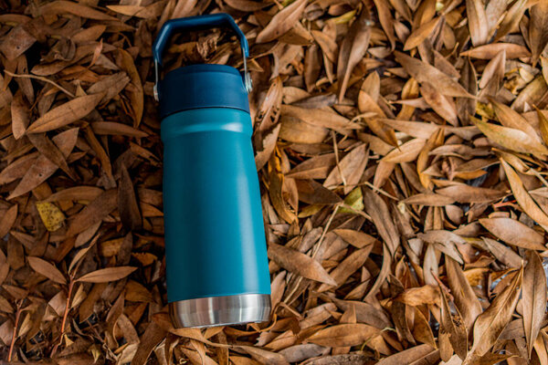 a blue thermos overturned on a pile of dry tree leaves. the thermos is one liter and has a handle.