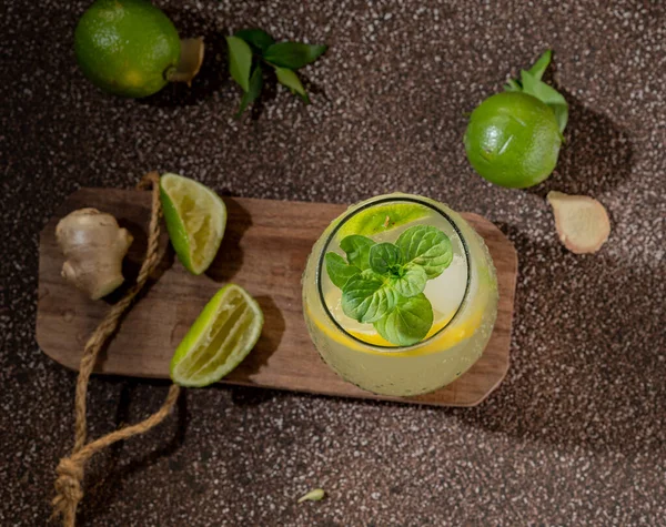 top view of a few green limes, some slices of the fruit and a few pieces of ginger, on a wooden board. There is a glass with lime juice, it has a slice of lemon that you can see.