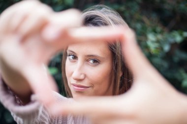 Kadın çerçeveleme hareketi yapıyor ve dışarıdaki kameraya bakıyor.