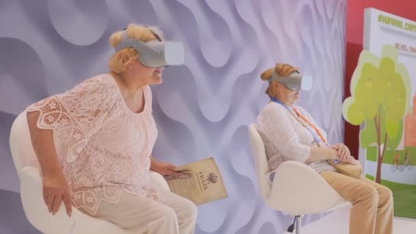Dwie starsze kobiety korzystające z zestawu słuchawkowego wirtualnej rzeczywistości na wystawie technologii VR — Wideo stockowe