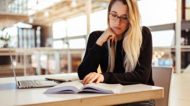 Kolej kütüphanesindeki masada diz üstü bilgisayar okuyarak sarı saçlı ve çene üzerine odaklanmış genç bir kız öğrenci.