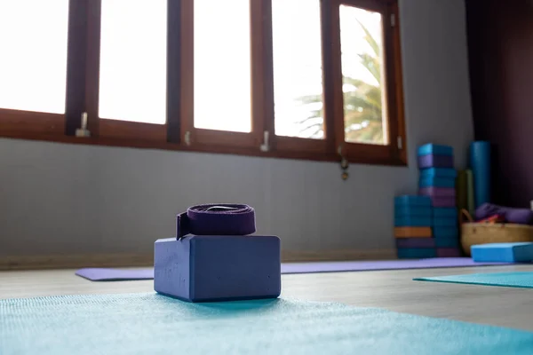Close-up of purple yoga block and belt with blue exercise mat placed on floor against windows in yoga studio