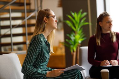 Young caucasian female coworkers with documents sitting on chairs and listening in meeting at corporate office