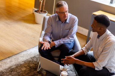 High angle view of multiracial mature businessman briefing project to young associate over laptop in meeting at corporate office