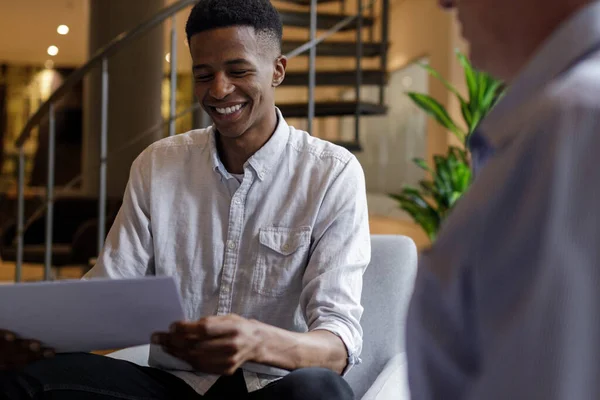 Smiling african american young businessman giving project report to caucasian mature partner while sitting in modern office