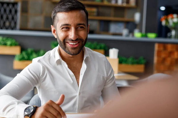 Smiling bearded young middle eastern businessman listening and looking at coworker while sitting in office cafe