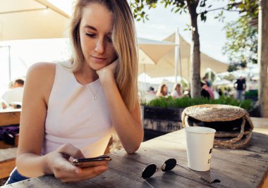 Beautiful blond woman with hand on chin using mobile phone while sitting by coffee cup and sunglasses at wooden table