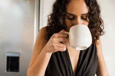 Close-up of mixed race beautiful woman with curly hair enjoying hot fresh coffee in kitchen during morning